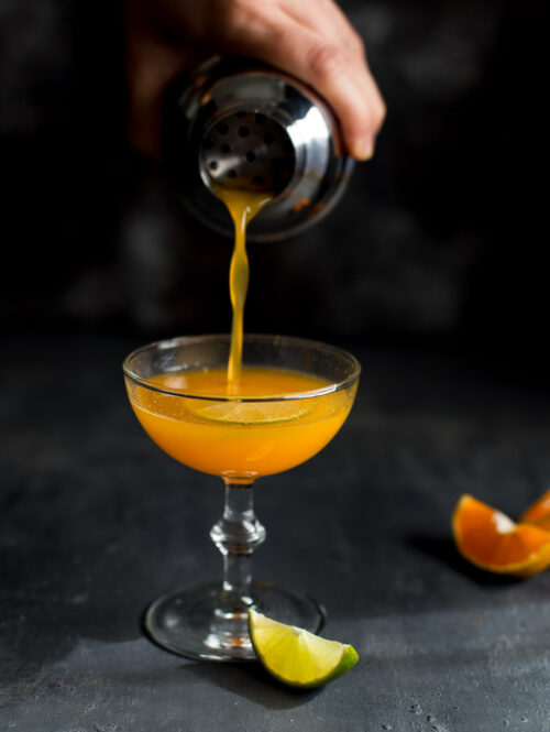 Orange vodka cocktail being poured from a shaker into a coupe glass.