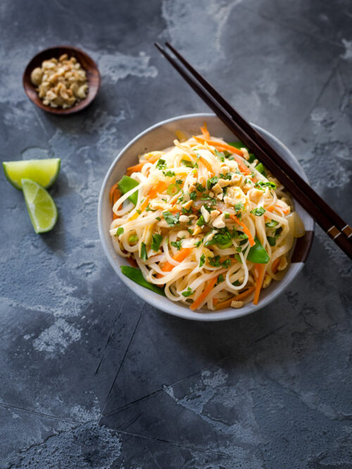 A bowl of chicken and veggie pad thai next to some lime wedges and a small dish of chopped peanuts.