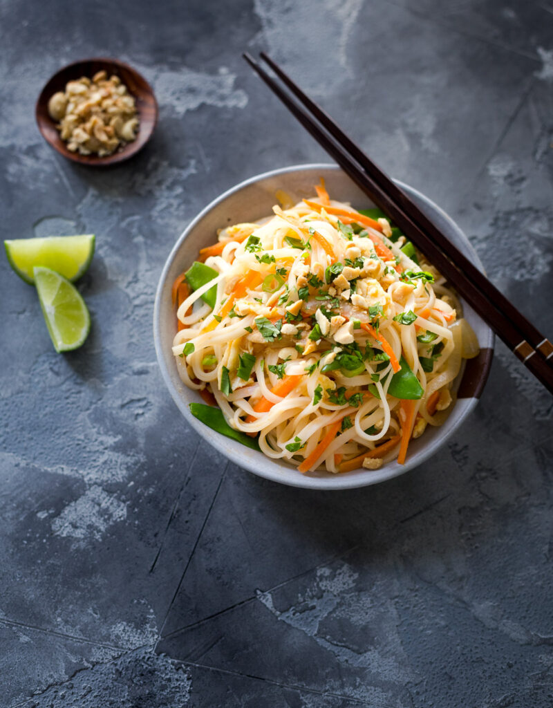 A bowl of chicken and veggie pad thai next to some lime wedges and a small dish of chopped peanuts.