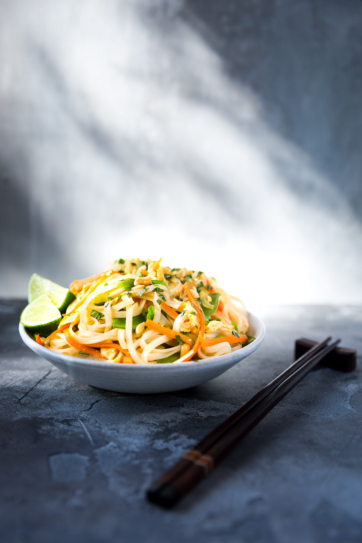 A bowl of chicken and veggie pad thai next to some chopsticks.