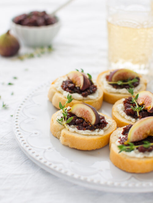 Fig and goat cheese crostini on a white plate with a glass of white wine in the background.
