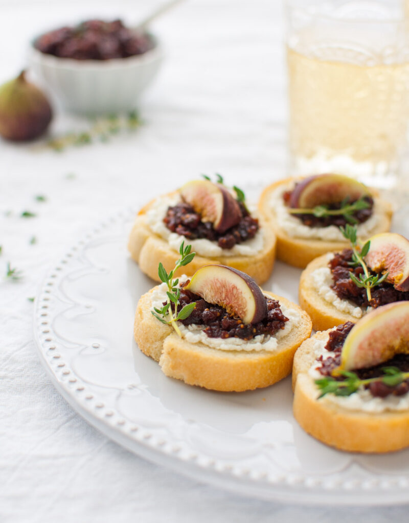 Fig and goat cheese crostini on a white plate with a glass of white wine in the background.