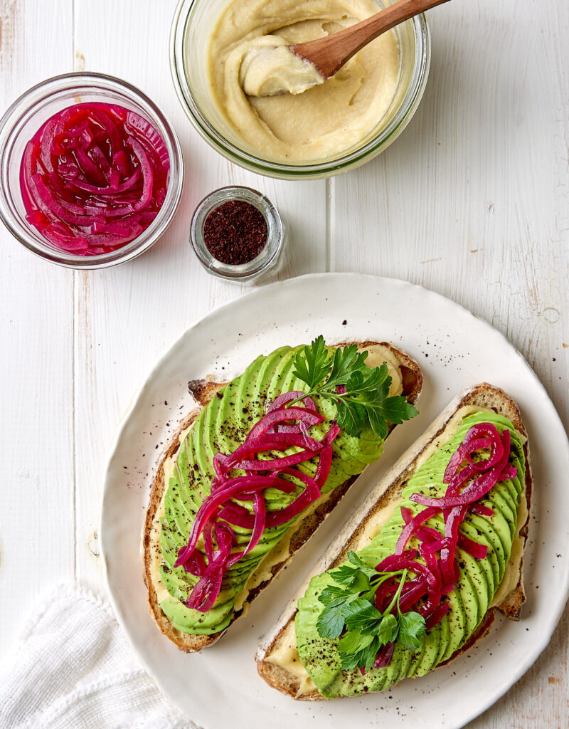 Two slices of avocado toast on a white plate next to some cashew cream and pickled onions.