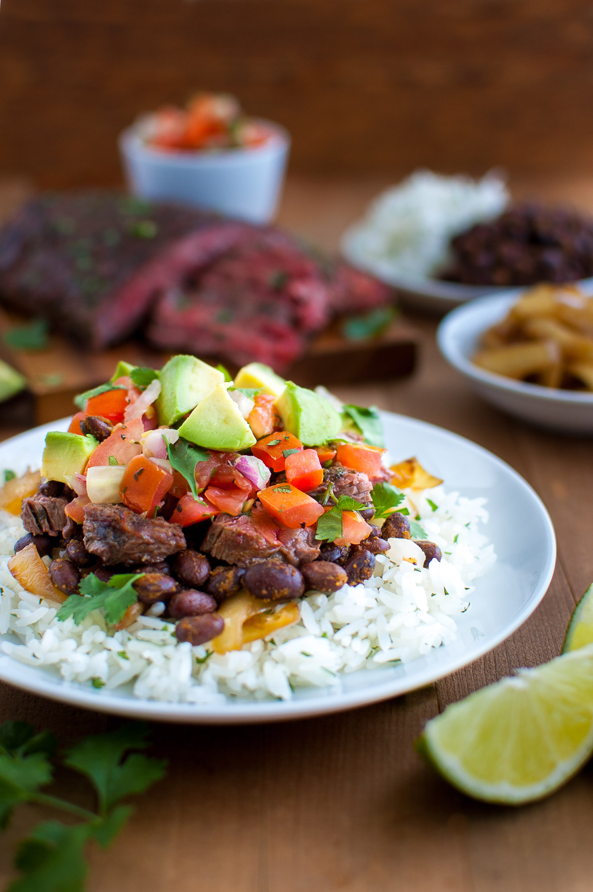A plate of rice topped with black beans, chopped avocados, pico de gallo, and cut steak.
