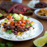 A plate of rice topped with black beans, chopped avocados, pico de gallo, and cut steak.