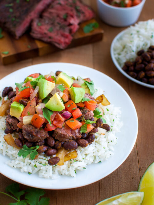 A plate of rice, black beans, chopped avocados, pico de gallo, and cut steak with more sliced steak in the background.