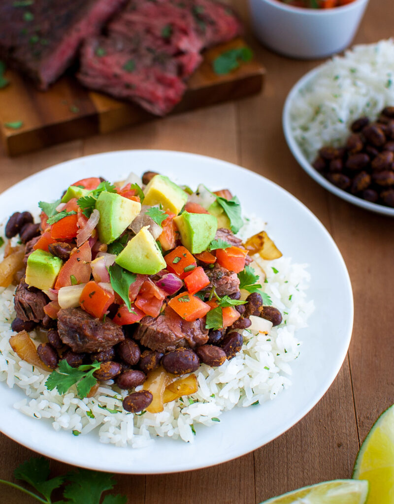 A plate of rice, black beans, chopped avocados, pico de gallo, and cut steak with more sliced steak in the background.