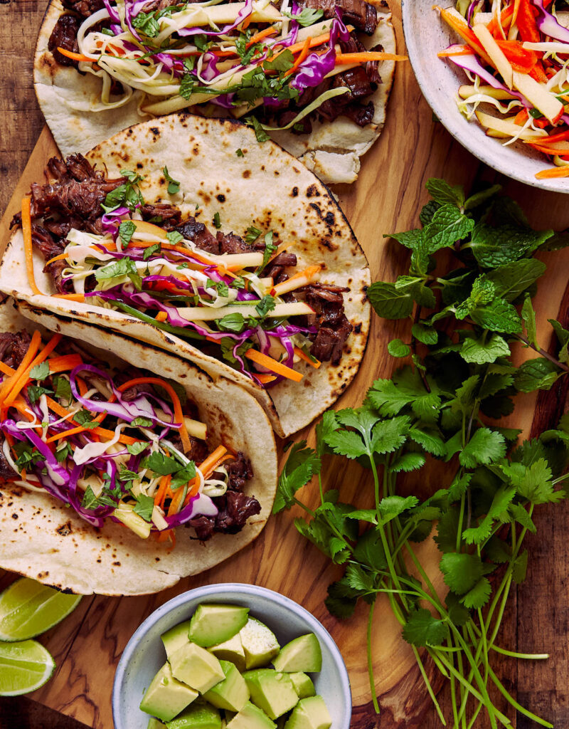 Beef tacos next to avocados, cabbage slaw, and cilantro.