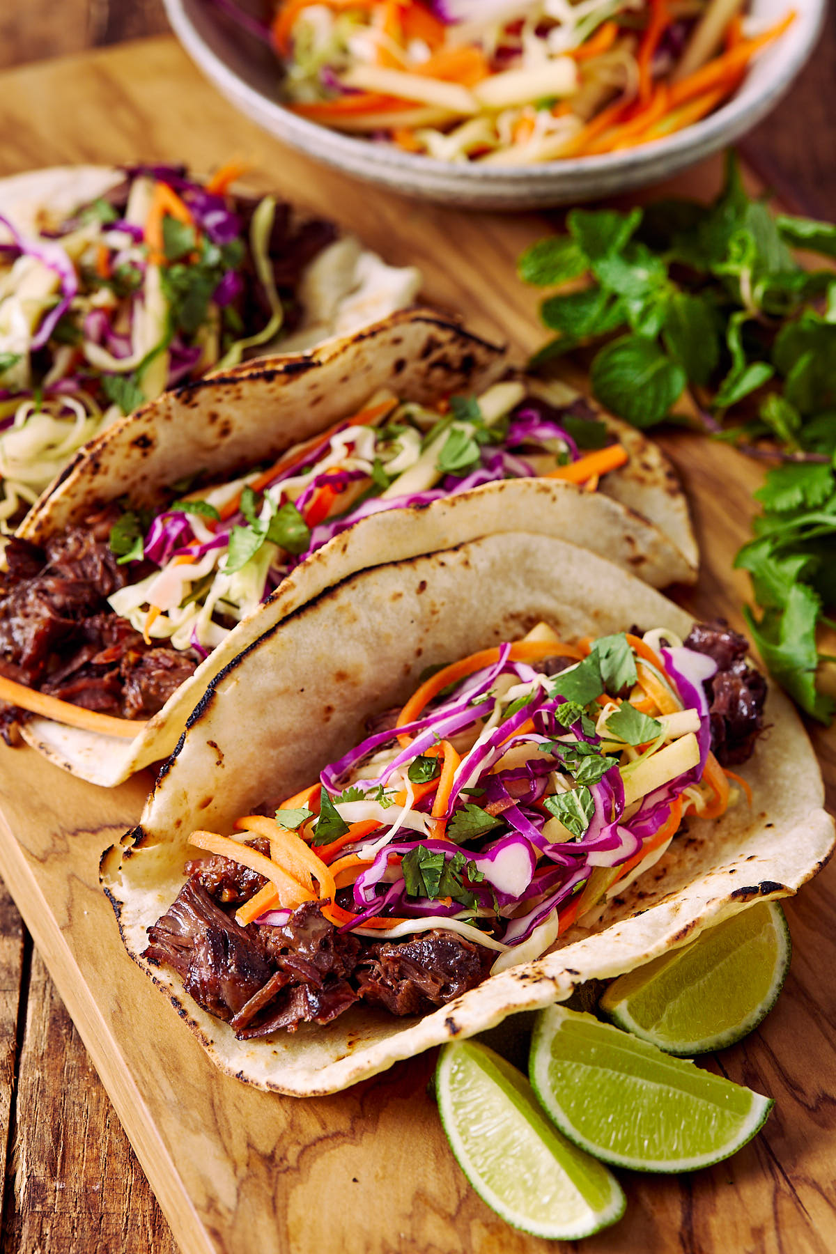 Three tacos on a wooden board with a bowl of slaw and some lime wedges.
