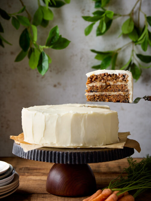 A slice of carrot cake being held up above a cake stand.