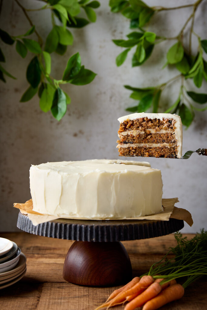 A slice of carrot cake being held up above a cake stand.