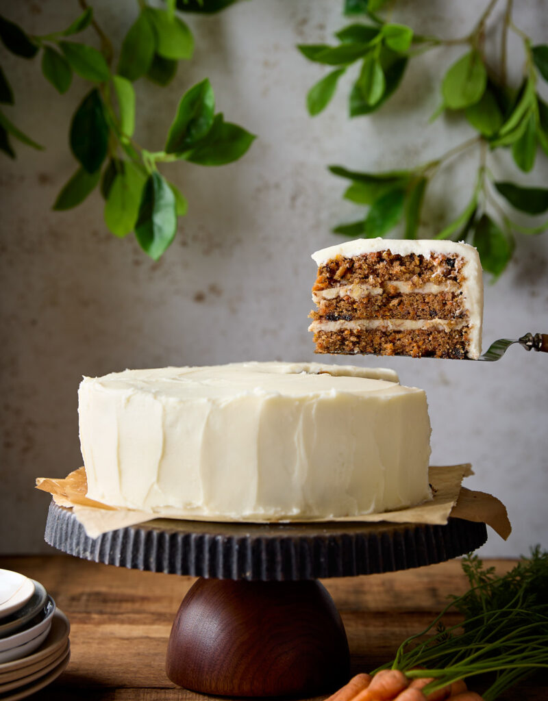 A slice of carrot cake being held up above a cake stand.